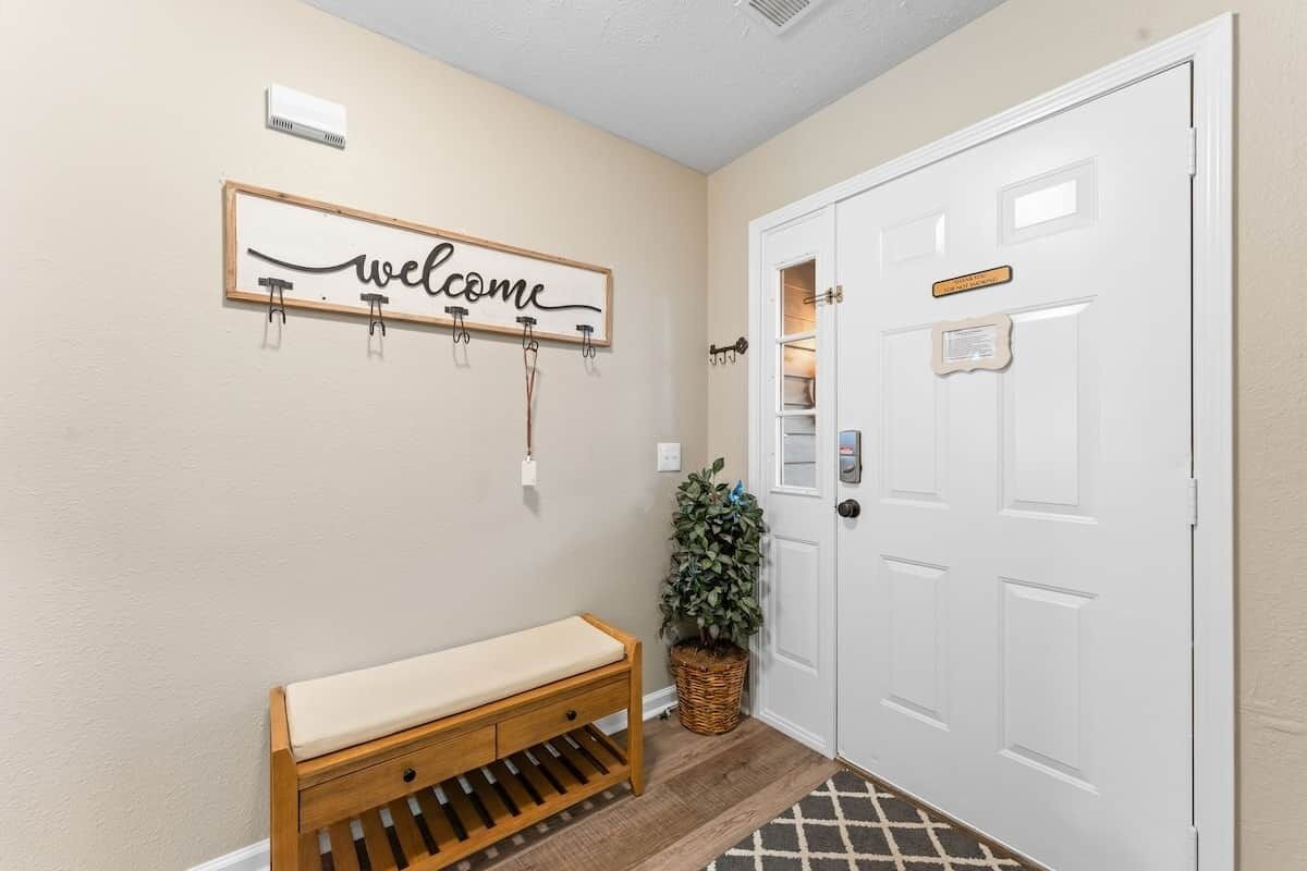 A hallway with a wooden bench, a welcome sign, a potted plant, and a white door.