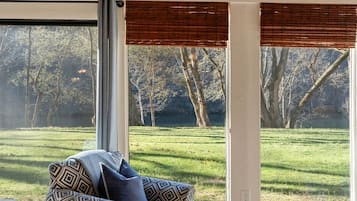 Ein Zimmer mit großem Fenster, einem gemusterten Sofa und Blick auf eine grüne Fläche mit Bäumen.