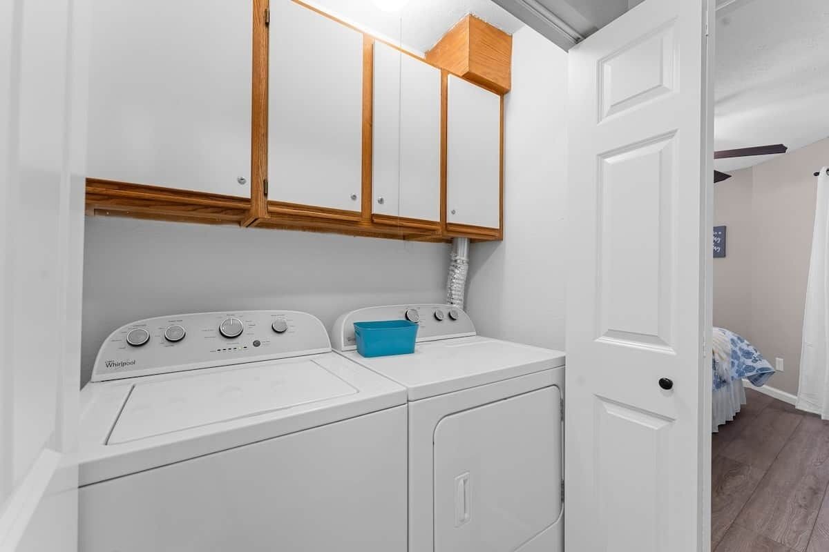 Un espace buanderie équipé d’un lave-linge et d’un sèche-linge à chargement frontal, de placards en bois au-dessus, et d’une porte donnant sur une autre pièce.