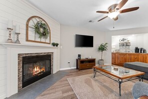 A living room with a fireplace, a flat-screen TV, a ceiling fan, and a kitchen area with a coffee station.