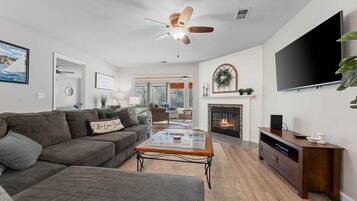 A living room with a sectional sofa, a glass coffee table, a fireplace, a TV, and a ceiling fan.