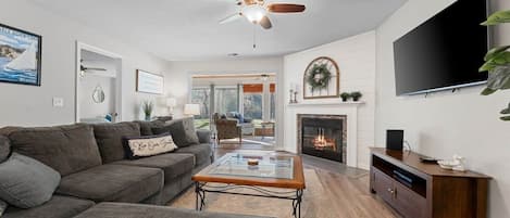A living room with a sectional sofa, a glass coffee table, a fireplace, a TV, and a ceiling fan.