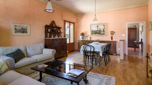 Ein Wohnzimmer mit Sofa, Couchtisch und Essbereich mit Stühlen und Tisch. Es gibt eine Kommode mit Dekorationselementen und ein Bild an der Wand.