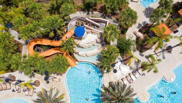Vista aérea de un resort con una gran piscina, tobogán acuático y zonas de descanso rodeadas de palmeras.