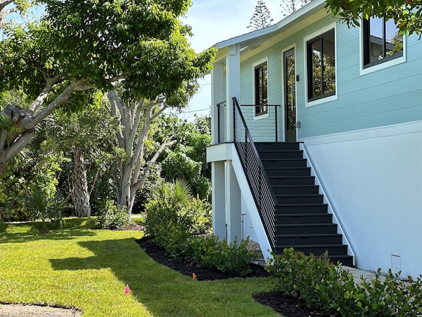A two-story house with a staircase leading to the entrance, surrounded by lush greenery and trees. - Camino Del Mar - Steps from the Beach! (Sanibel)