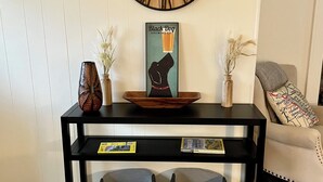Une pièce avec un plancher en bois, une table noire sur laquelle se trouvent une horloge, un vase et un tableau encadré.