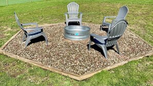 Un foyer entouré de trois chaises Adirondack, installé sur une surface de gravier avec de l’herbe en arrière-plan.