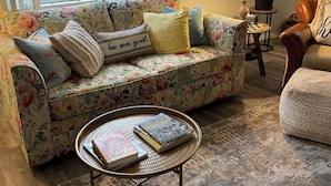 Un salon chaleureux avec un canapé à motifs floraux, une table basse ronde sur laquelle sont posés des livres, une lampe et une plante en pot.