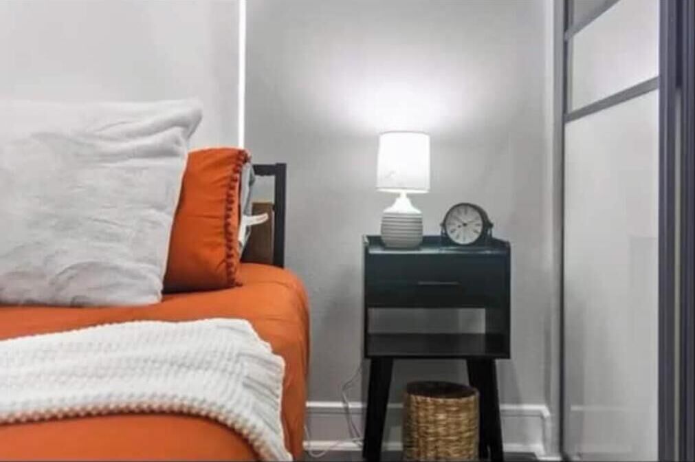 Un dormitorio con una cama, una mesita de noche con lámpara, un reloj y una cesta.