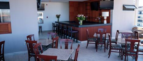 A hotel dining area with wooden tables and chairs, a bar with a TV, and a view of the city.