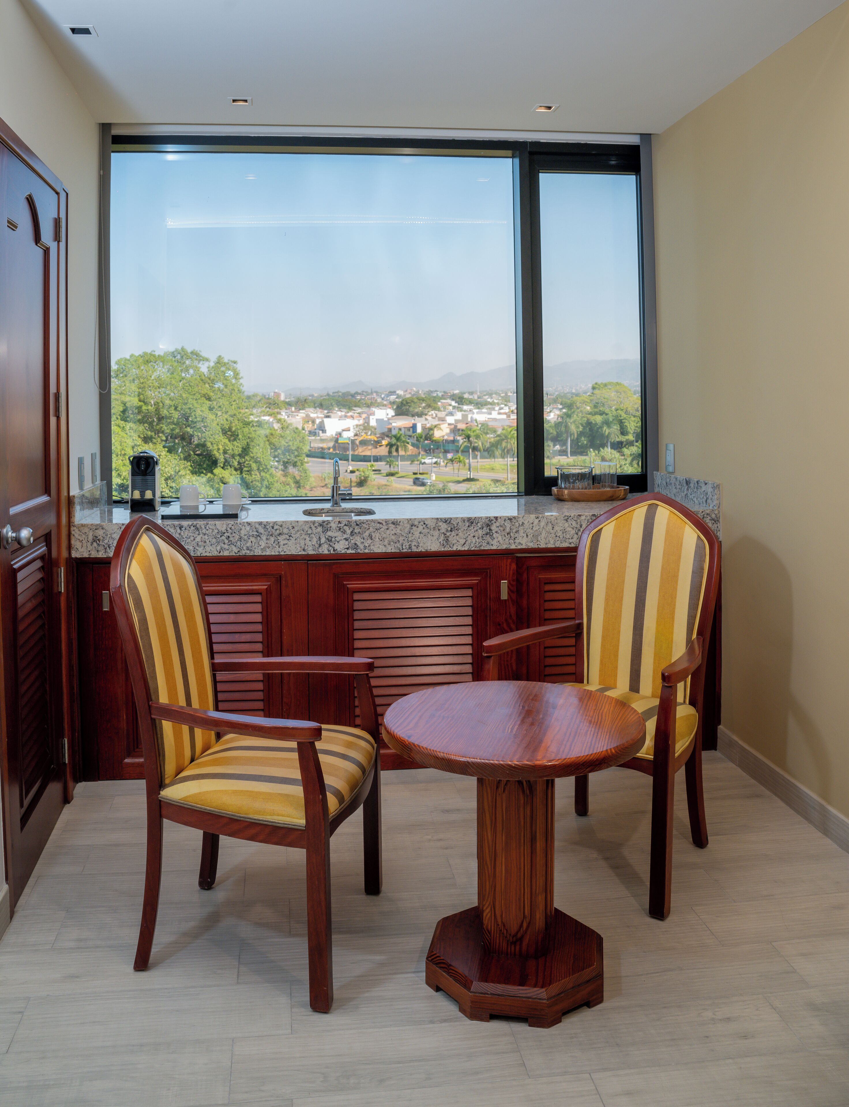 Un comedor con una mesa redonda de madera y dos sillas, una ventana con vistas a un paisaje urbano, y una encimera de cocina con una cafetera y vasos.