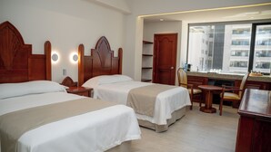 Habitación de hotel con dos camas, una zona de estar con mesa y sillas, y un ventanal con vistas a la ciudad.