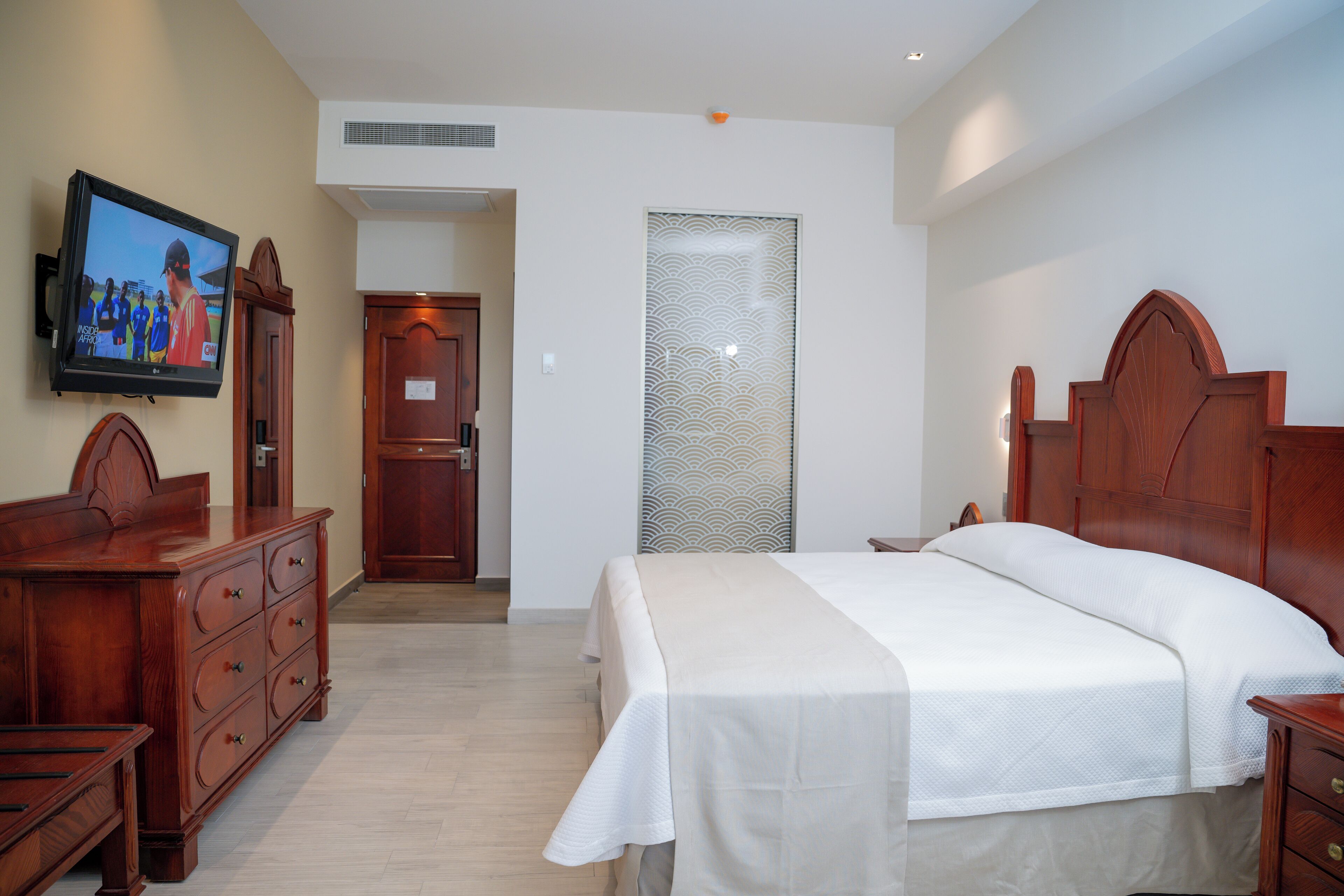 Une chambre d’hôtel avec un lit en bois, un téléviseur fixé au mur, une commode en bois et une porte donnant sur une autre pièce.