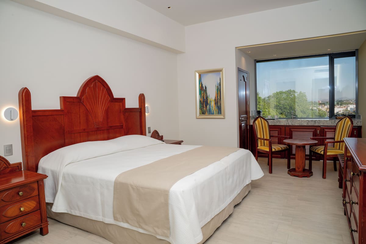 Quarto de hotel com cama grande, cabeceira de madeira, mesas de cabeceira, escrivaninha com cadeiras e uma janela ampla com vista para a cidade.