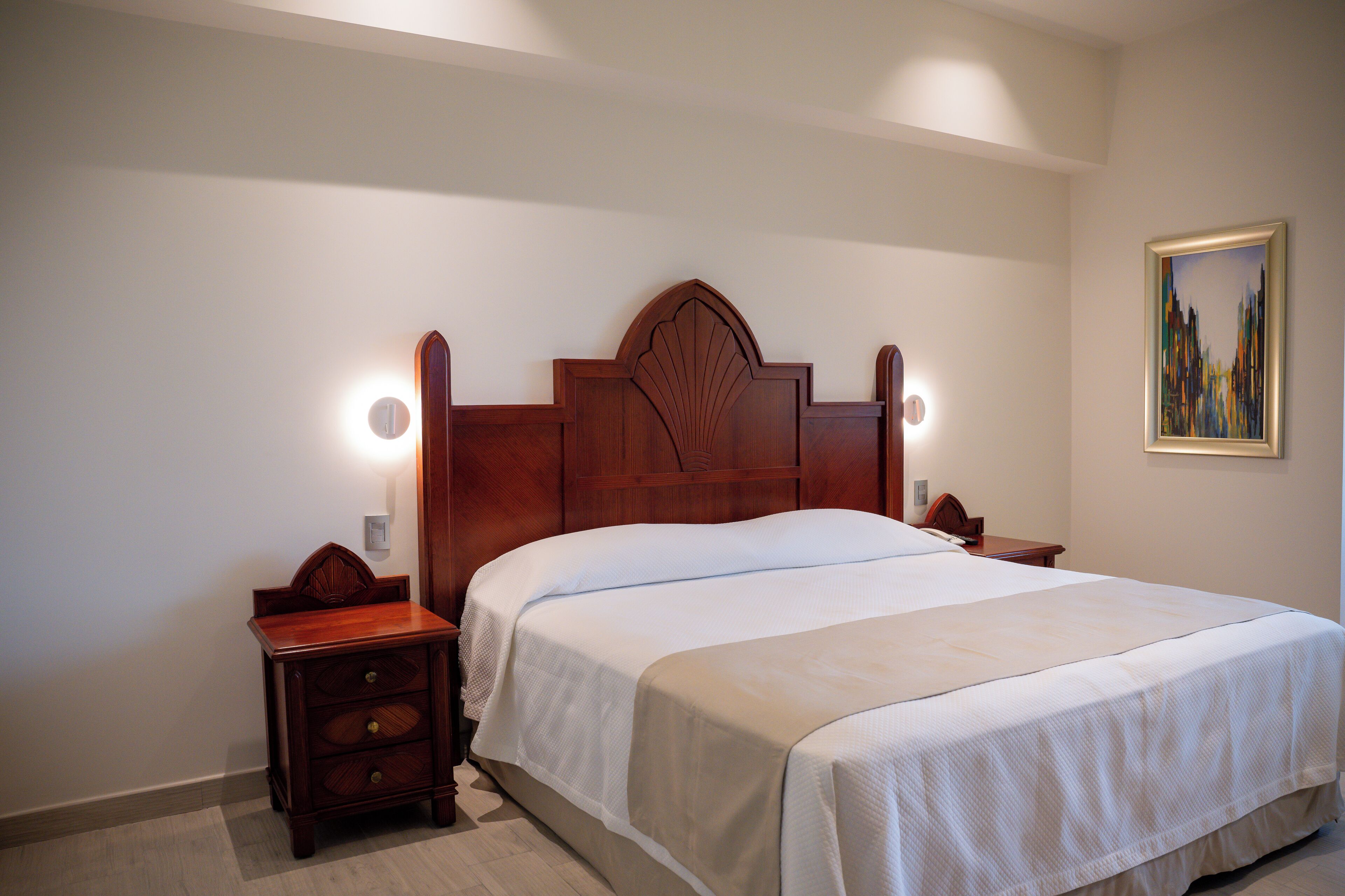 A neatly made bed with a wooden headboard, a nightstand with drawers, and a wall-mounted lamp.