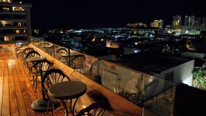 Un bar en la azotea con suelo de madera y barandillas de cristal, que ofrece vistas a un paisaje urbano nocturno.