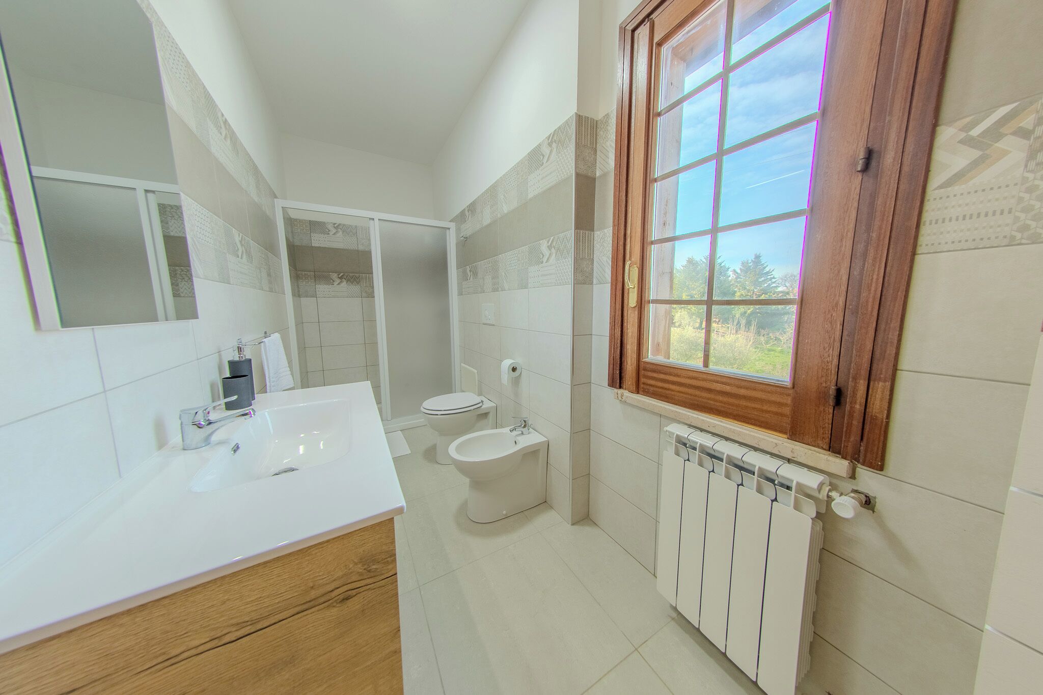 A modern bathroom with a white sink, toilet, and bidet, a large mirror, and a window with a view of greenery.