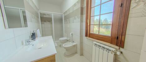 A modern bathroom with a white sink, toilet, and bidet, a large mirror, and a window with a view of greenery.
