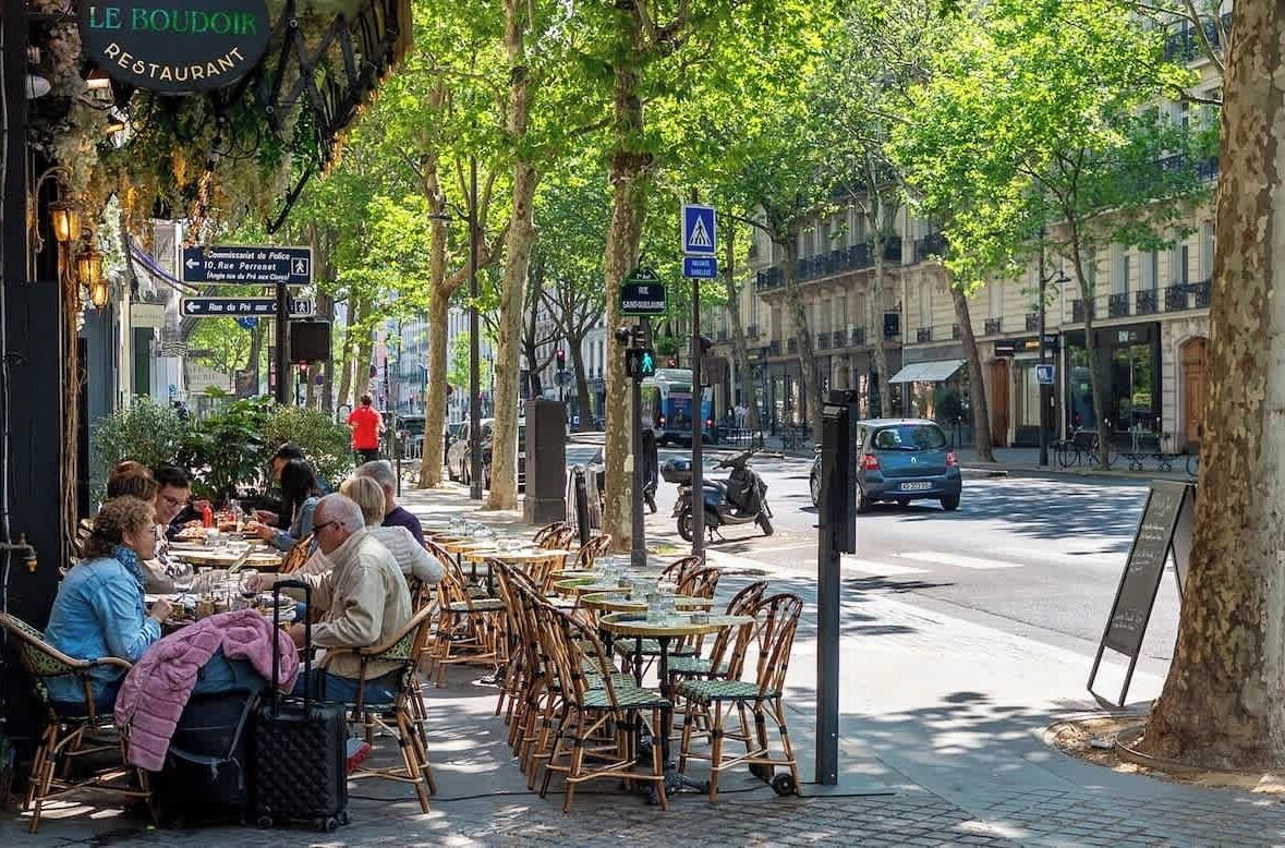 Outdoor seating with tables and chairs, a street view with cars and a pedestrian, and a sign for 'LE BOUDOIR RESTAURANT'.