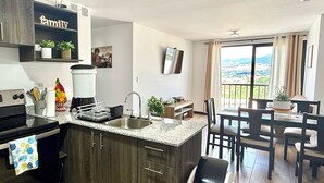 Una cocina moderna con armarios de madera oscura, encimera de granito y un lavamanos integrado. Hay un comedor con mesa y sillas, y vistas al exterior a través de un amplio ventanal.