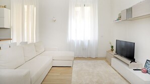 A modern living room with a white sofa, a flat-screen TV on a stand, a small side table with a plant, and sheer curtains on the windows. - Corsica Apartment Travel Inside  (MILANO)