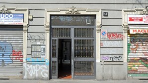 A building entrance with a metal gate, a closed shutter on the left, and a shop with a sign on the right. - Corsica Apartment Travel Inside  (MILANO)