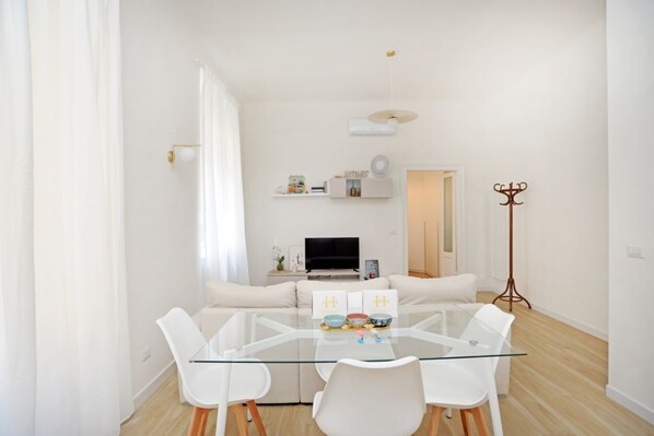 A modern living room with a glass dining table, white sofas, a TV, and a coat rack.