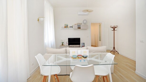 A modern living room with a glass dining table, white sofas, a TV, and a coat rack. - Corsica Apartment Travel Inside  (MILANO)