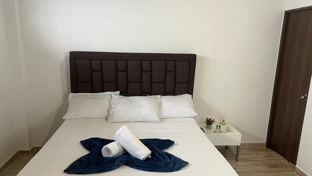 A neatly made bed with white linens and a dark blue blanket, a small bedside table with a plant, and a wooden door in a simple room. - HOTEL SARAPE ESTADIO (Medellín)