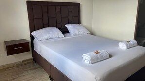 A neatly made bed with white linens and two rolled towels, a dark headboard, and a small wooden nightstand. - HOTEL SARAPE ESTADIO (Medellín)