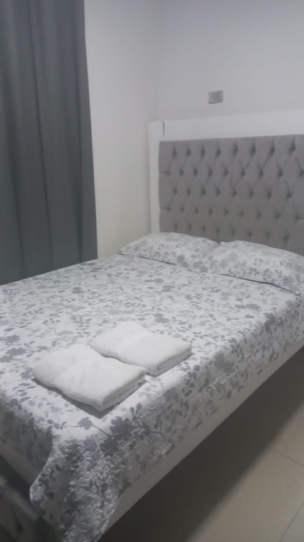 A bed with a floral-patterned bedspread and two pillows, a grey headboard, and a grey curtain.