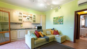 Un salon avec un canapé beige, des coussins colorés, un coin cuisine avec des meubles de couleur claire, et un tableau accroché au mur.