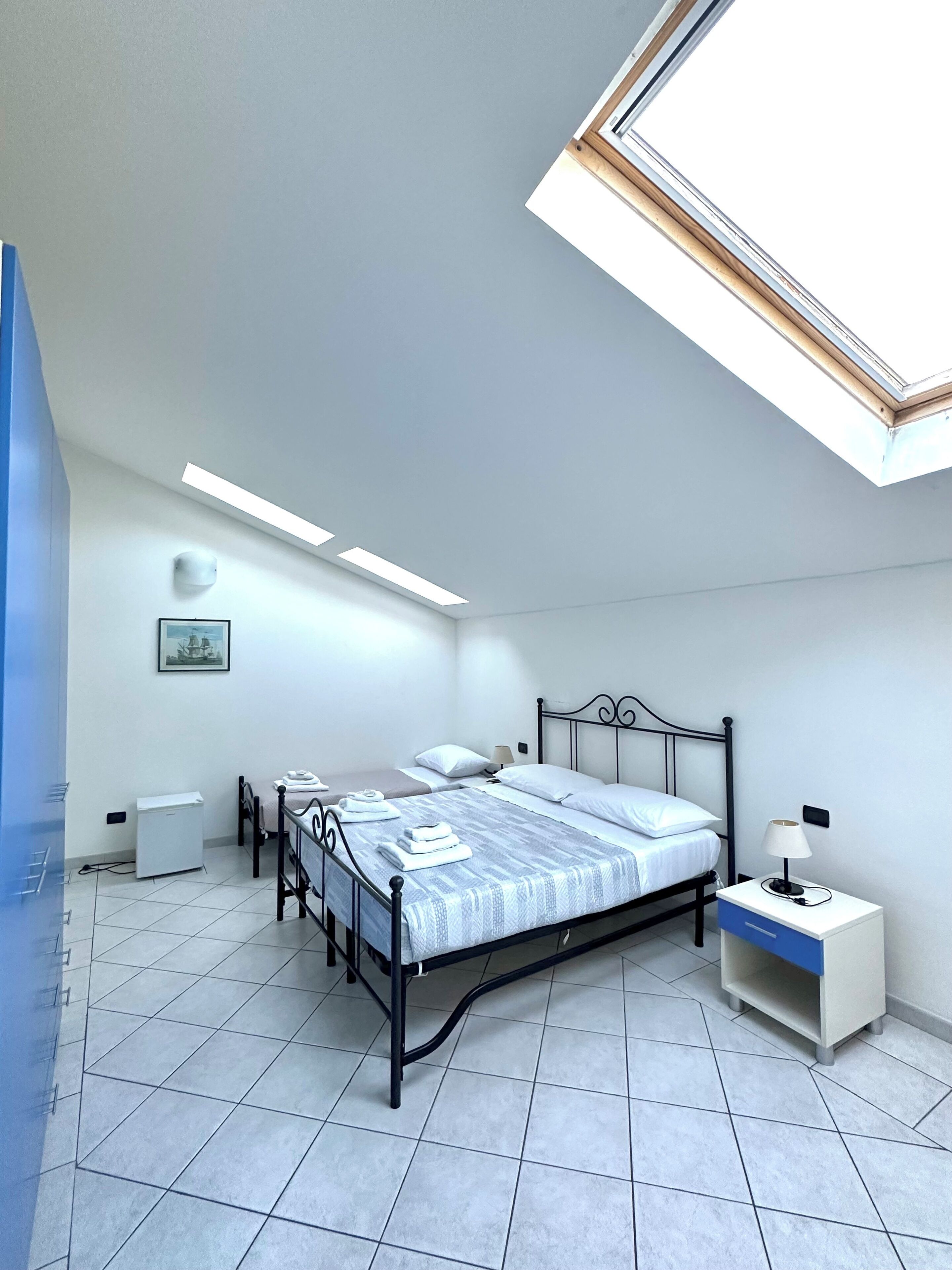 Un dormitorio con tragaluces, una cama con cabecera, una mesita de noche con lámpara y un pequeño armario blanco.