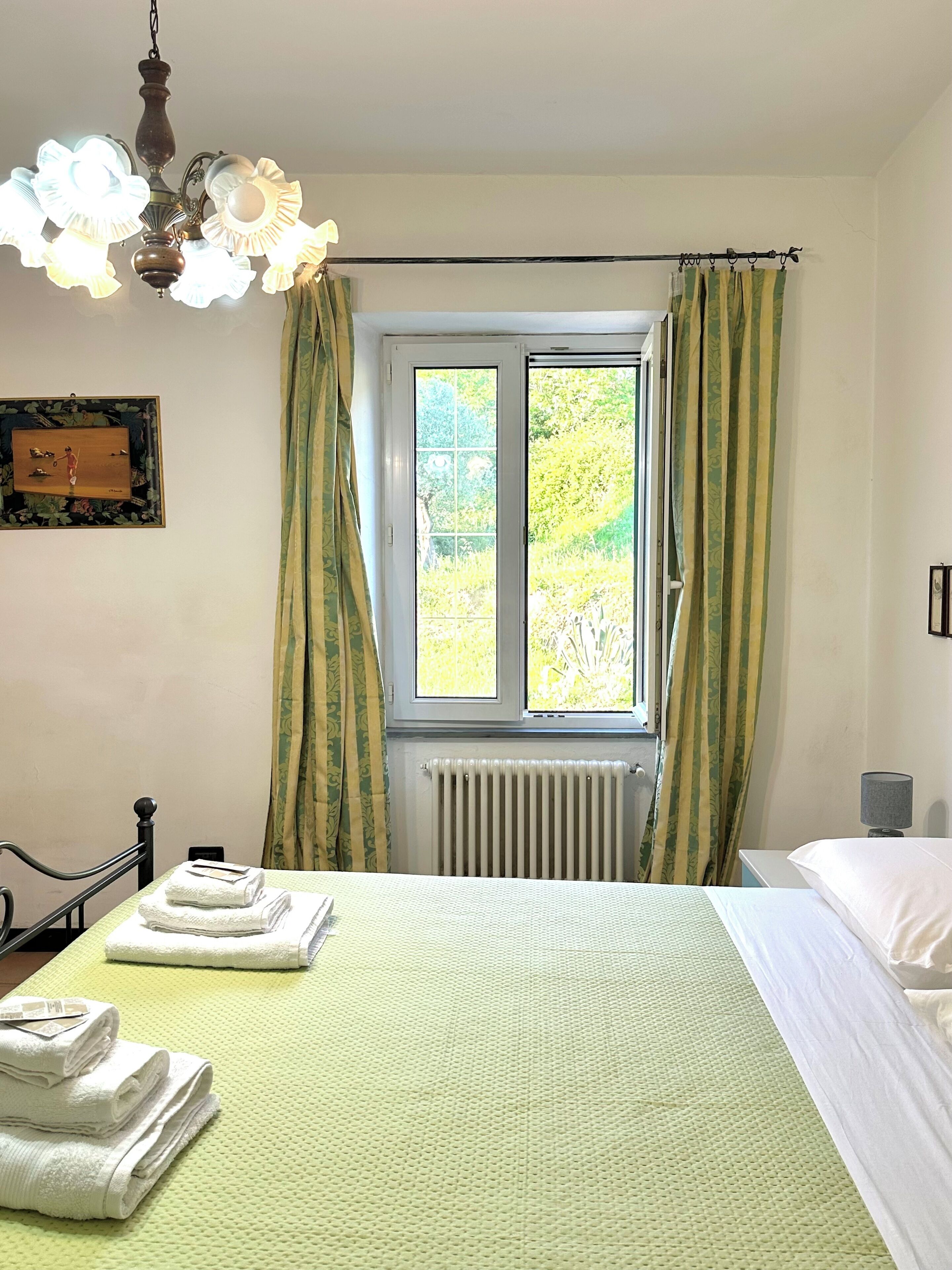 Un dormitorio con cama, radiador, ventana con cortinas, lámpara colgante y un cuadro en la pared.