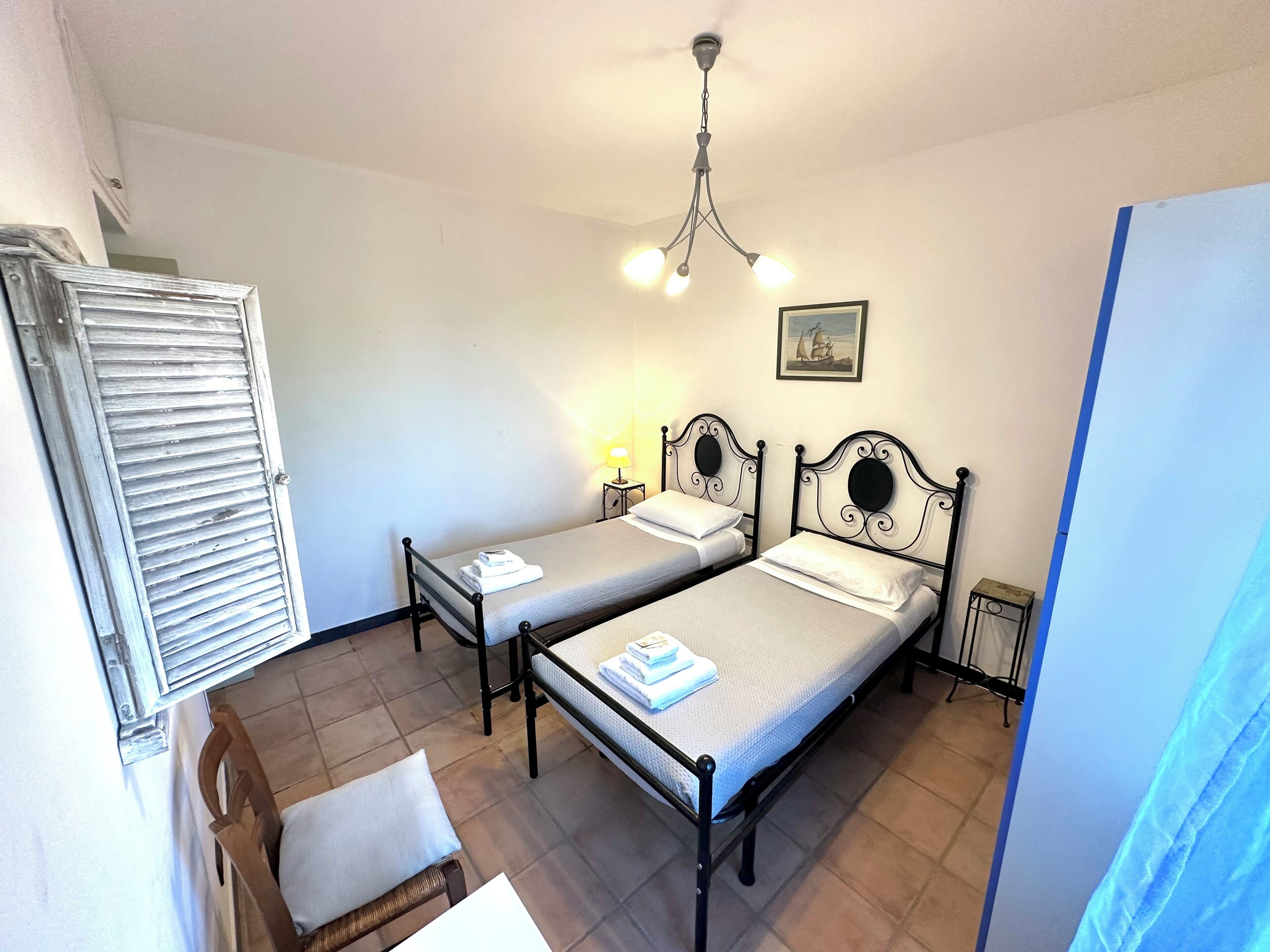 A room with two single beds, a wooden chair, a small table with a lamp, and a ceiling fan.