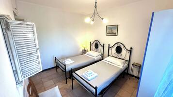 A room with two single beds, a wooden chair, a small table with a lamp, and a ceiling fan.