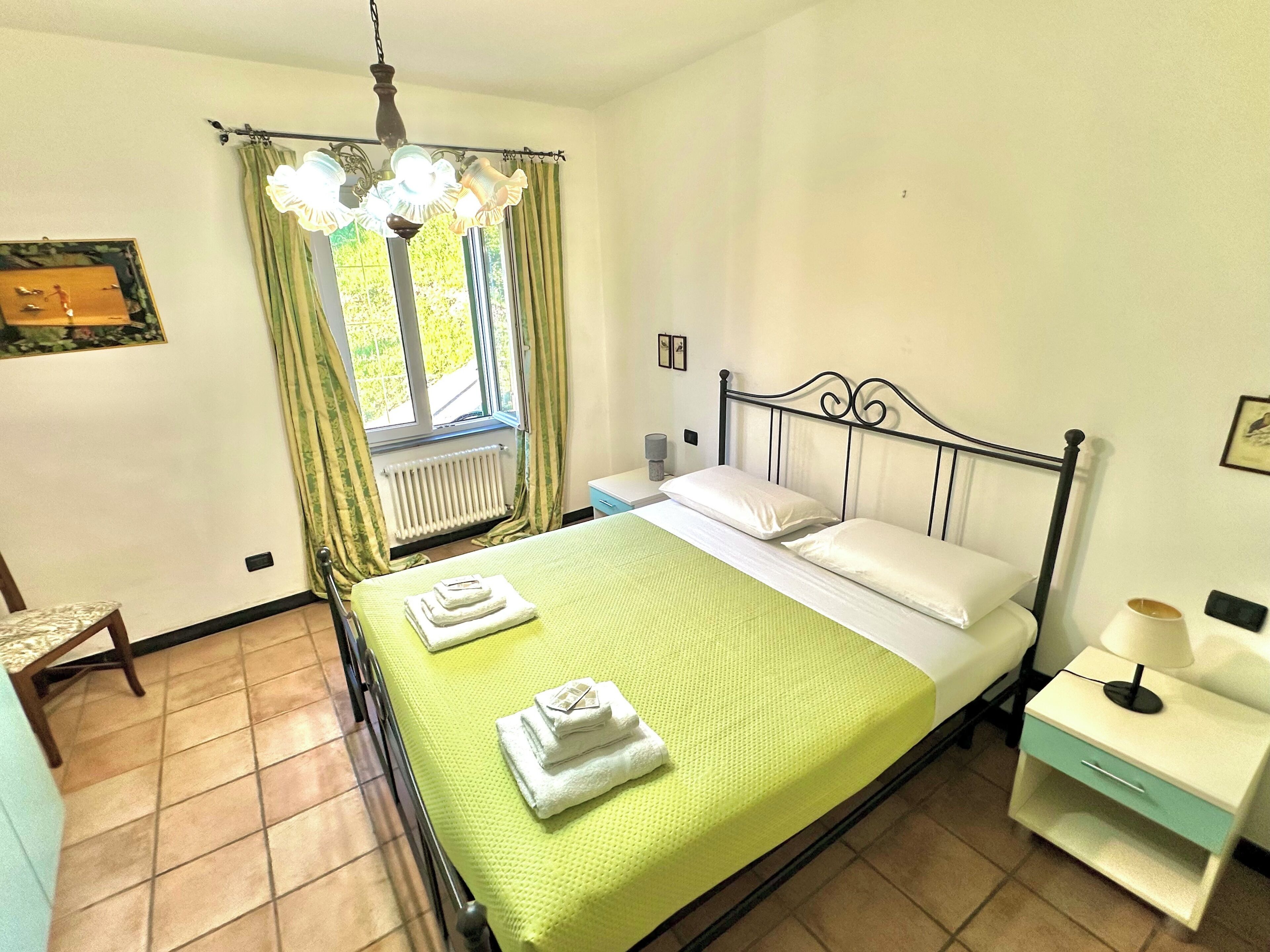 A neatly made bed with a green bedspread, two pillows, and folded towels. There is a nightstand with a lamp and a small drawer. A window with curtains and a radiator is visible, along with a chair and a framed picture on the wall.