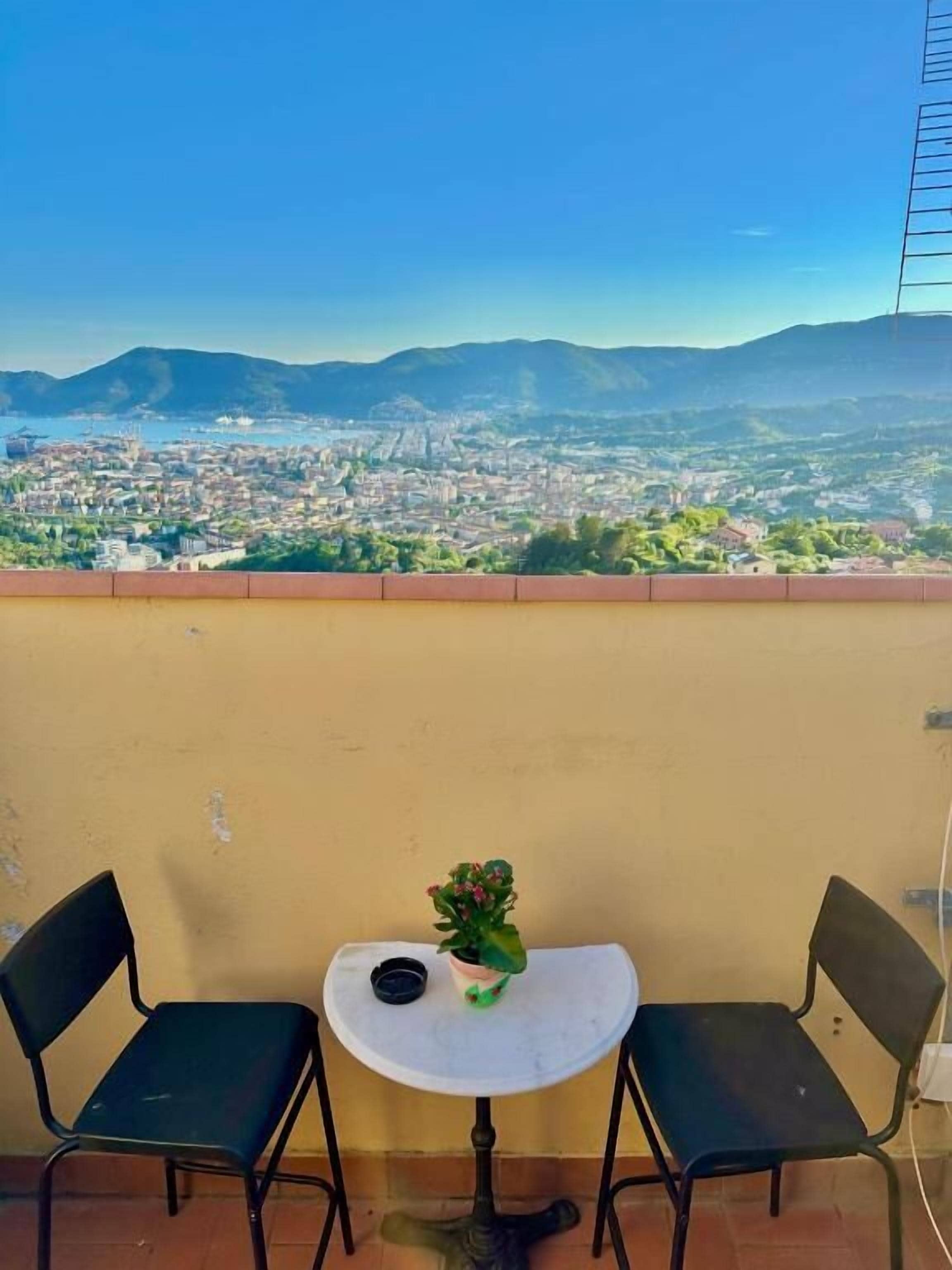 A balcony with two chairs and a small table, overlooking a cityscape with mountains in the distance.