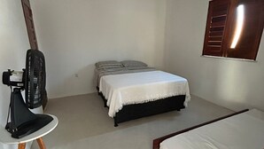 A small, sparsely furnished room with a single bed, a small table with a fan, and a window with wooden shutters. - Casa da Paz  (Trairi)