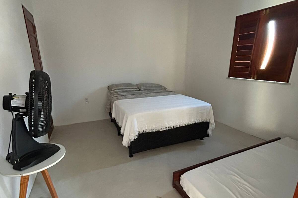 A small, sparsely furnished room with a single bed, a small table with a fan, and a window with wooden shutters.