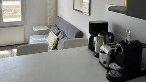 A modern, well-lit room with a grey sofa, a small table, a coffee maker, and a toaster. There is a window with a view of the outside, and an air conditioning unit on the wall. - Nice T2 on the little port of Sanary, Clim (Sanary-sur-Mer)