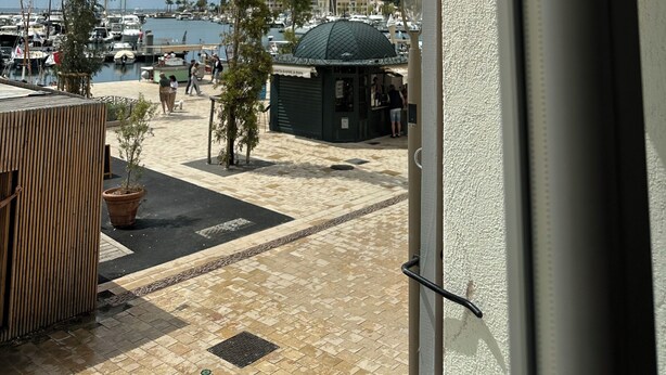 A view from a balcony overlooking a marina with boats, a small kiosk, and a tiled patio area. - Nice T2 on the little port of Sanary, Clim (Sanary-sur-Mer)