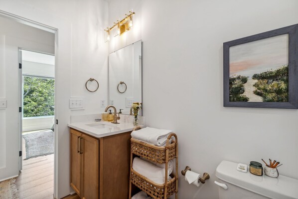 A bathroom with a white vanity, a wicker towel rack, a framed painting, and a toilet with a small shelf.