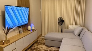 A modern living room with a sectional sofa, a flat-screen TV, a standing fan, and a large window with curtains. - Glamorous 2BR/1Bath/Wifi/Netflix/Heater/Washing/5starBnB (Pasay)