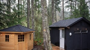 Two wooden cabins in a forest, one with a black roof and the other with a brown roof, both with windows and doors. - Wellness Retreat at Lake Cushman. Sauna, Cold Plunge, Fire Pit (Hoodsport)
