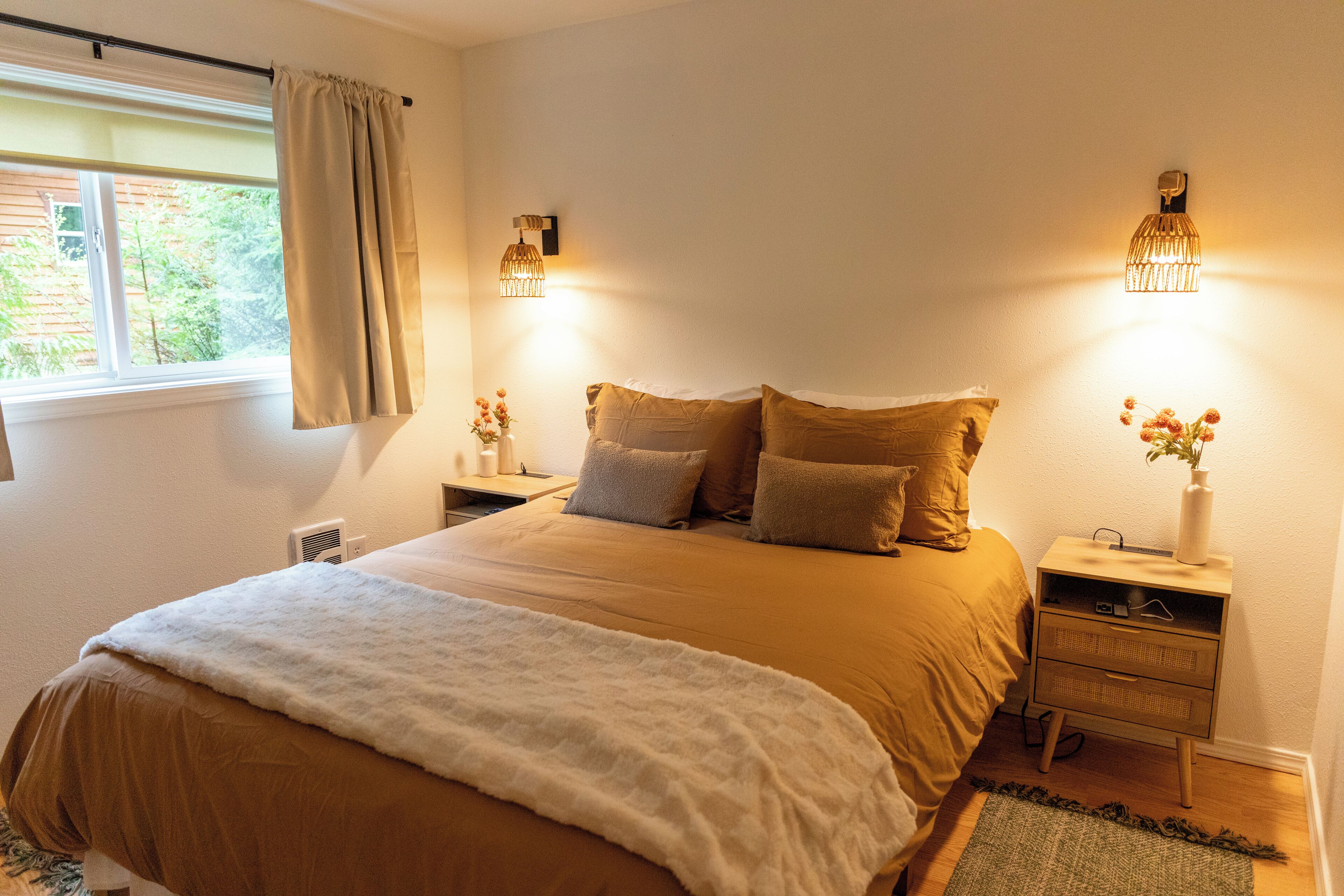 A bedroom with a large bed, two bedside tables, a nightstand, a window with curtains, and wall-mounted lights.