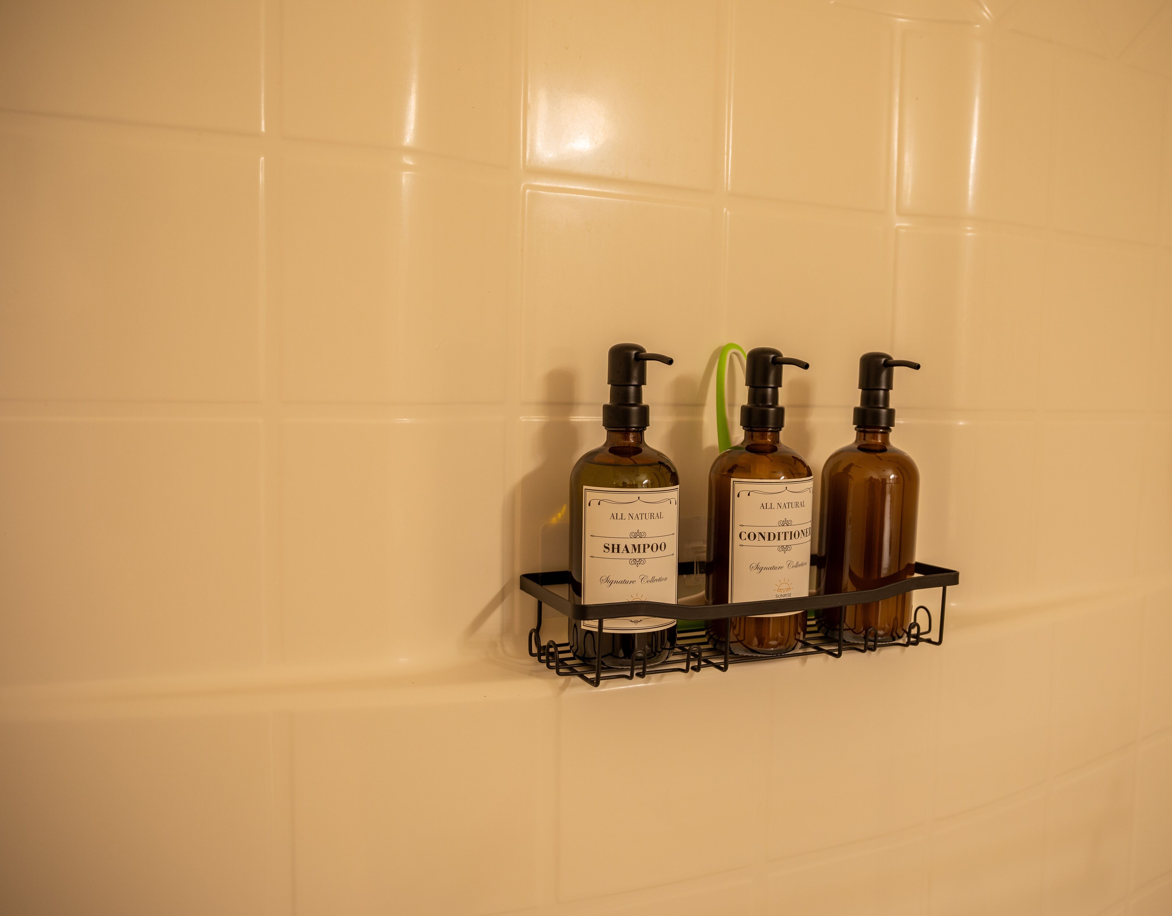 A bathroom with a tiled wall and a shelf holding three bottles of All Natural shampoo and conditioner.