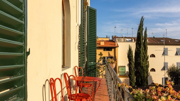 A balcony with red metal chairs and a table, overlooking a garden with plants and a building with a yellow facade and green shutters. - Modern, Bright and Spacious 2-Bedroom Apartment (Firenze)
