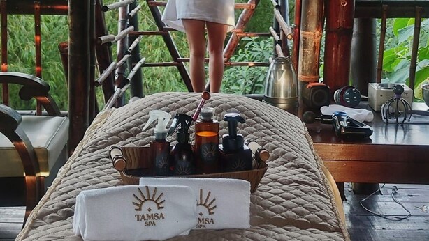 Iemand staat op een balkon met uitzicht op weelderige begroeiing, met een massagetafel op de voorgrond met een handdoek en flessen.