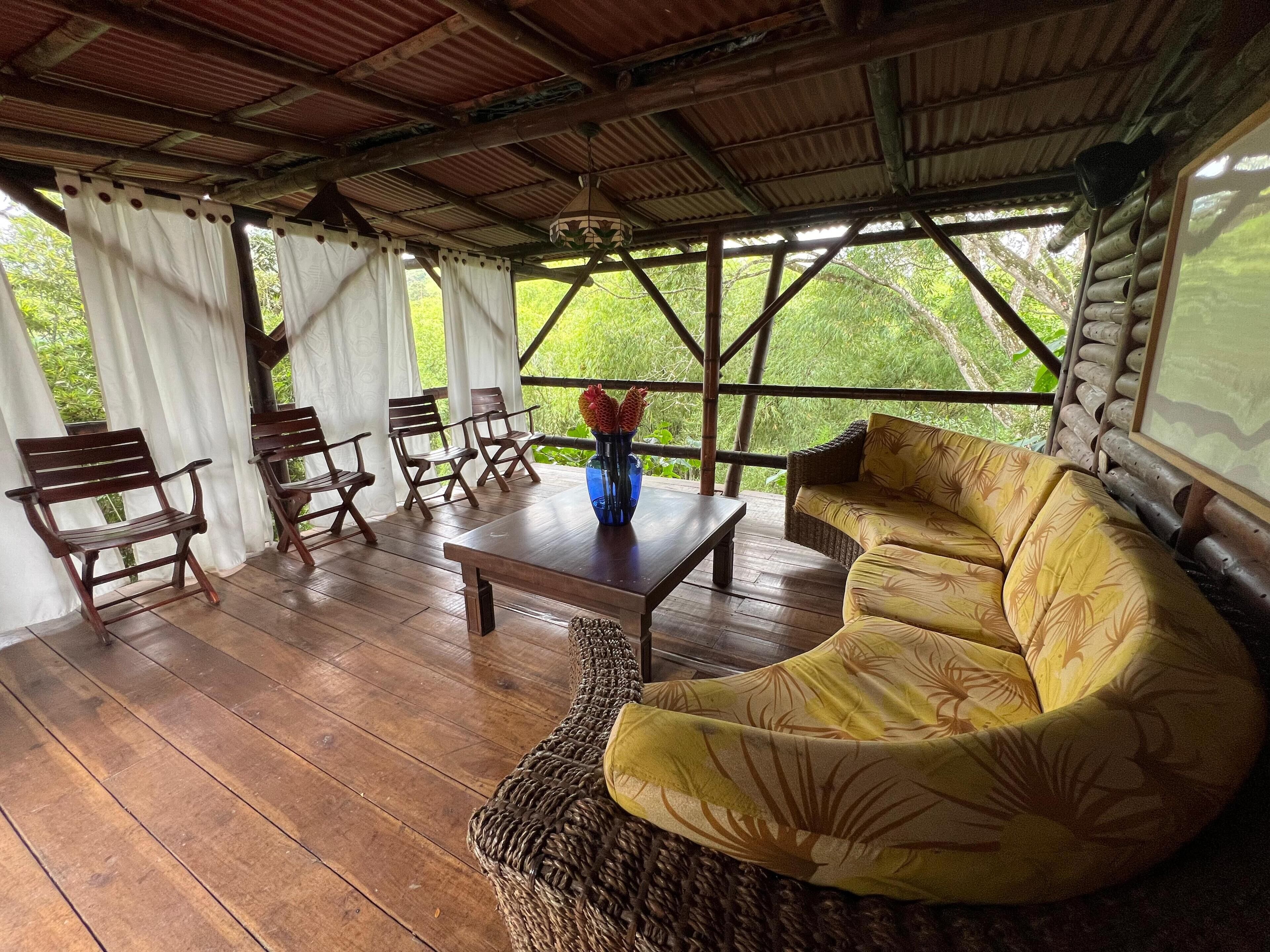 A covered outdoor seating area with wooden furniture and a woven sofa, featuring a view of greenery.